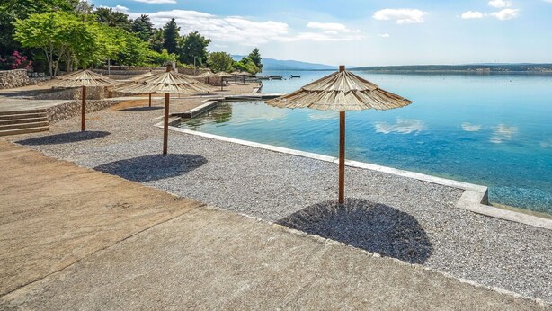 A poolside area with clear blue water, surrounded by pebble ground, and shaded by straw umbrellas.