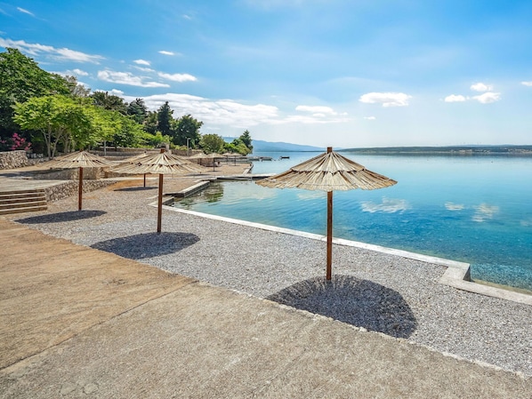A poolside area with clear blue water, surrounded by pebble ground and shaded by straw umbrellas.
