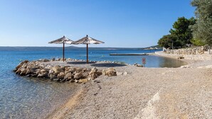 A pebble beach with two thatched umbrellas, a clear blue sea, and a stone wall with trees.