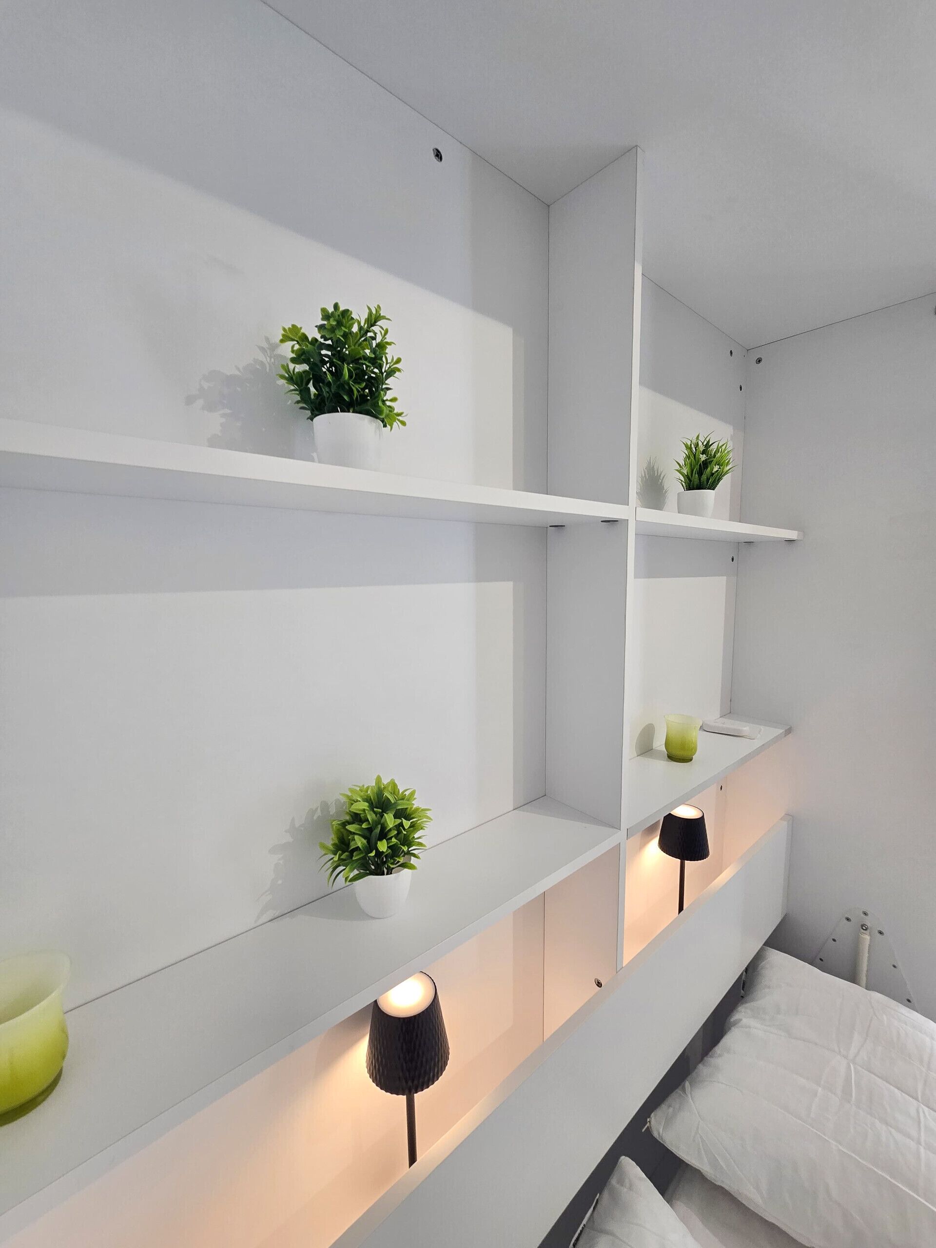 A modern bedroom with white walls, a bed with white bedding, and two bedside lamps. There are three white shelves with potted plants.