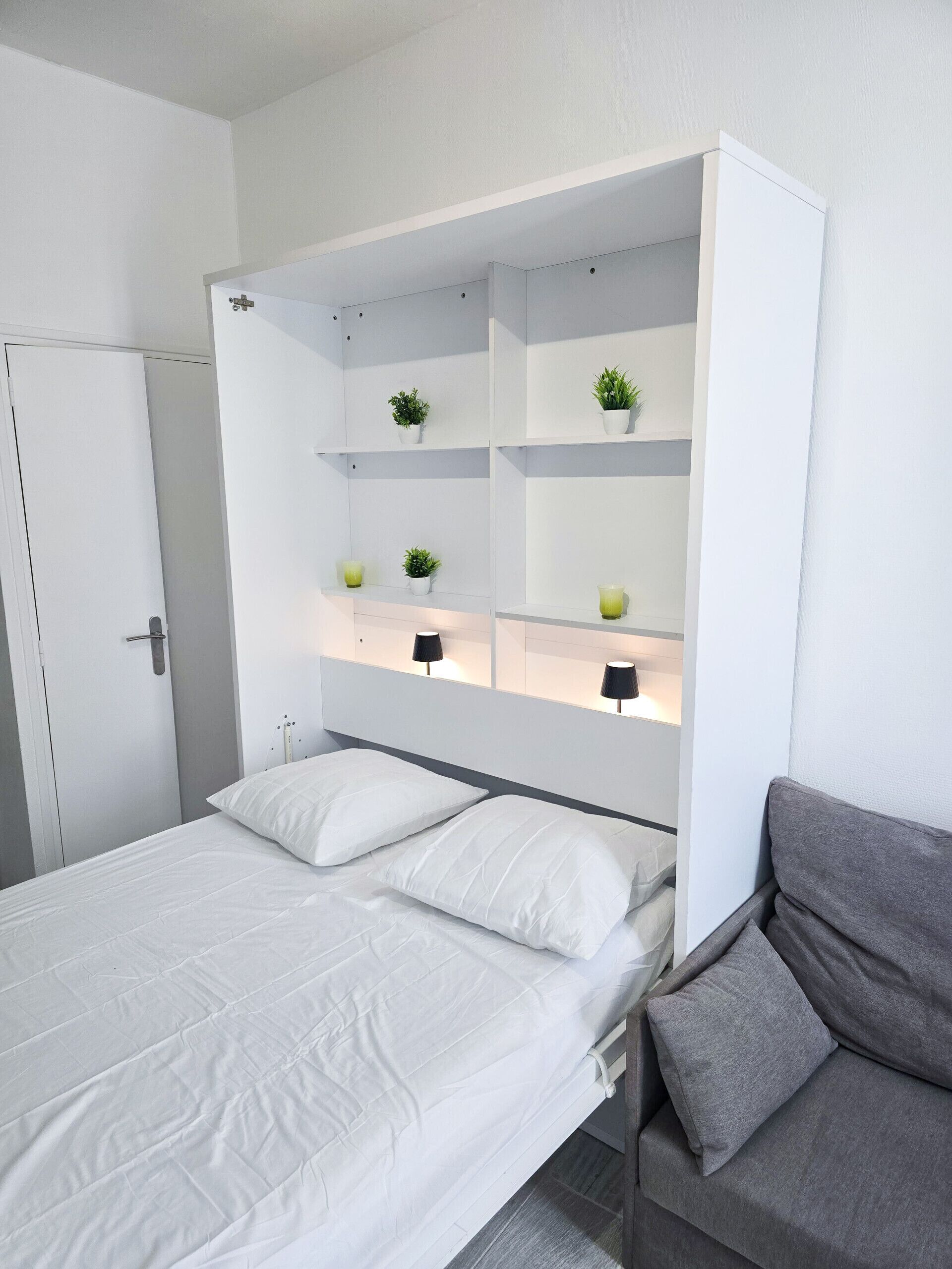 Een moderne slaapkamer met een bed, een grijze bank en ingebouwde kasten met planten en lampen.