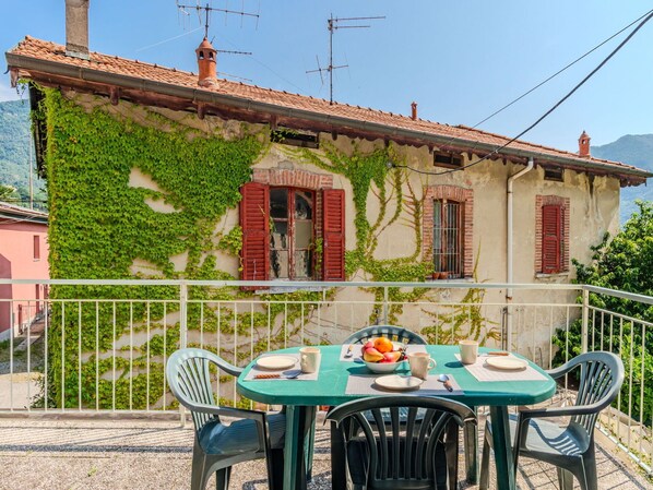 A table set with plates, cups, and a bowl of fruit on a balcony with a view of a building covered in ivy.