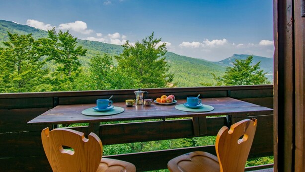 Una mesa de madera con dos sillas, un plato de fruta y dos tazas en un balcón con vistas a un paisaje montañoso.