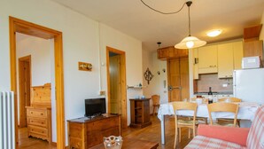 Una sala de estar acogedora con un sofá de madera, un televisor y una mesa de comedor con sillas. Se observa una zona de cocina con armarios y un refrigerador.