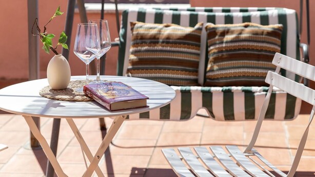 A round table with a book and two glasses, a small vase with a plant, and a striped sofa with pillows in the background. - Beautiful apartment for 2 guests with WIFI, A/C, TV, terrace and pets allowed (Olbia)