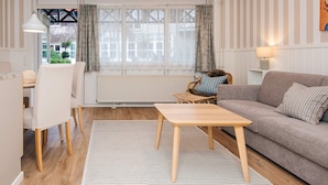 Ein modernes Wohnzimmer mit einer Couch, einem Esstisch und einem Sessel. Ein Fenster mit Vorhängen, ein Heizkörper und eine Lampe auf einem Beistelltisch sind zu sehen.
