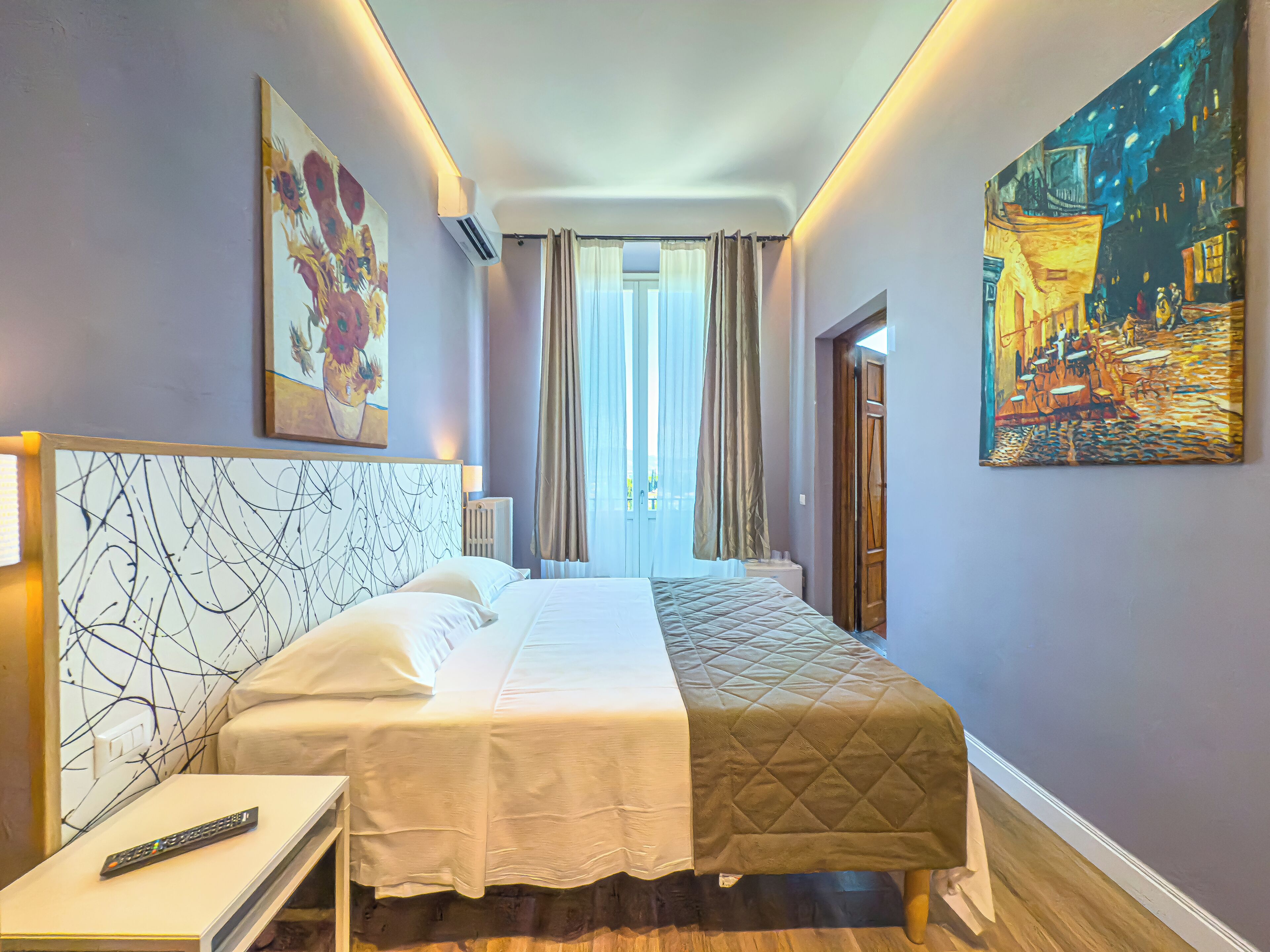A hotel room with a bed, a nightstand, a painting, and a view of the street through the window.
