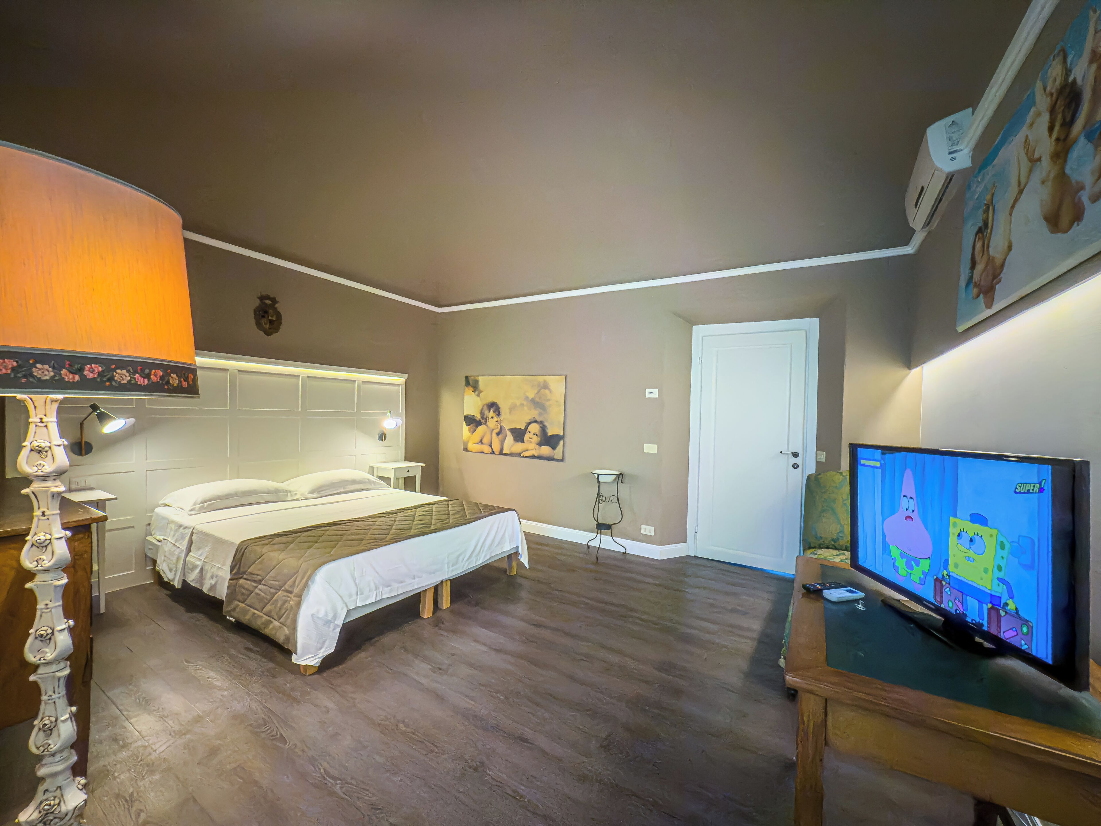 A hotel room with a bed, a nightstand, a lamp, a painting on the wall, a door, and a television displaying an animated show.