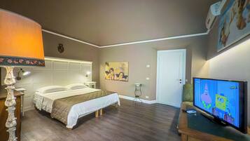 A hotel room with a bed, a nightstand, a lamp, a painting on the wall, a door, and a television displaying an animated show.