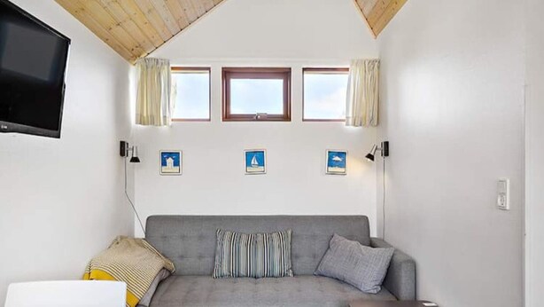 Een gezellige kamer met een grijze bank, een klein houten tafeltje en een aan de muur bevestigde televisie. Drie ramen met gordijnen en enkele kleine prenten op de muur.