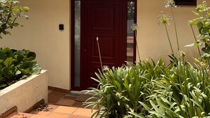 A building with a red-tiled roof, a red door, and a small window. There are plants in front of the door and a tiled patio area.