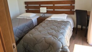 A room with two single beds, each with a wooden headboard and white pillows. There is a small table with a lamp between the beds.