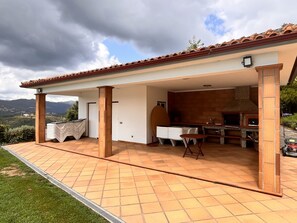 Une terrasse extérieure couverte, dotée d’une cheminée en briques, d’une table à manger avec des chaises et offrant une vue sur le paysage environnant.