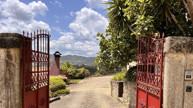 A cobblestone pathway leading to a gated entrance with a red iron gate, surrounded by greenery and a mountainous backdrop.