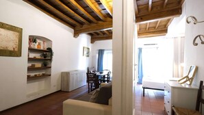 Ein Wohnzimmer mit Holzboden, einer Couch, einem Esstisch mit Stühlen und einer Küchenzeile mit Regalen und einem Heizkörper.