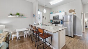 A modern kitchen with a central island, white cabinetry, stainless steel appliances, and a dining area with bar stools. - Dog-Friendly Coastal Retreat | Resort Pools & Walk to Beach (Corpus Christi)