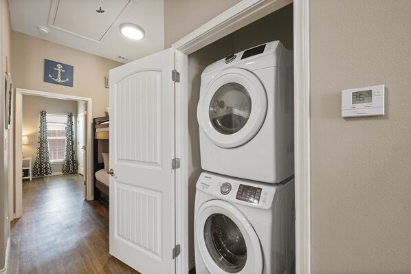A laundry room with stacked washers and dryers, a thermostat, and a door leading to another room. - Dog-Friendly Coastal Retreat | Resort Pools & Walk to Beach (Corpus Christi)