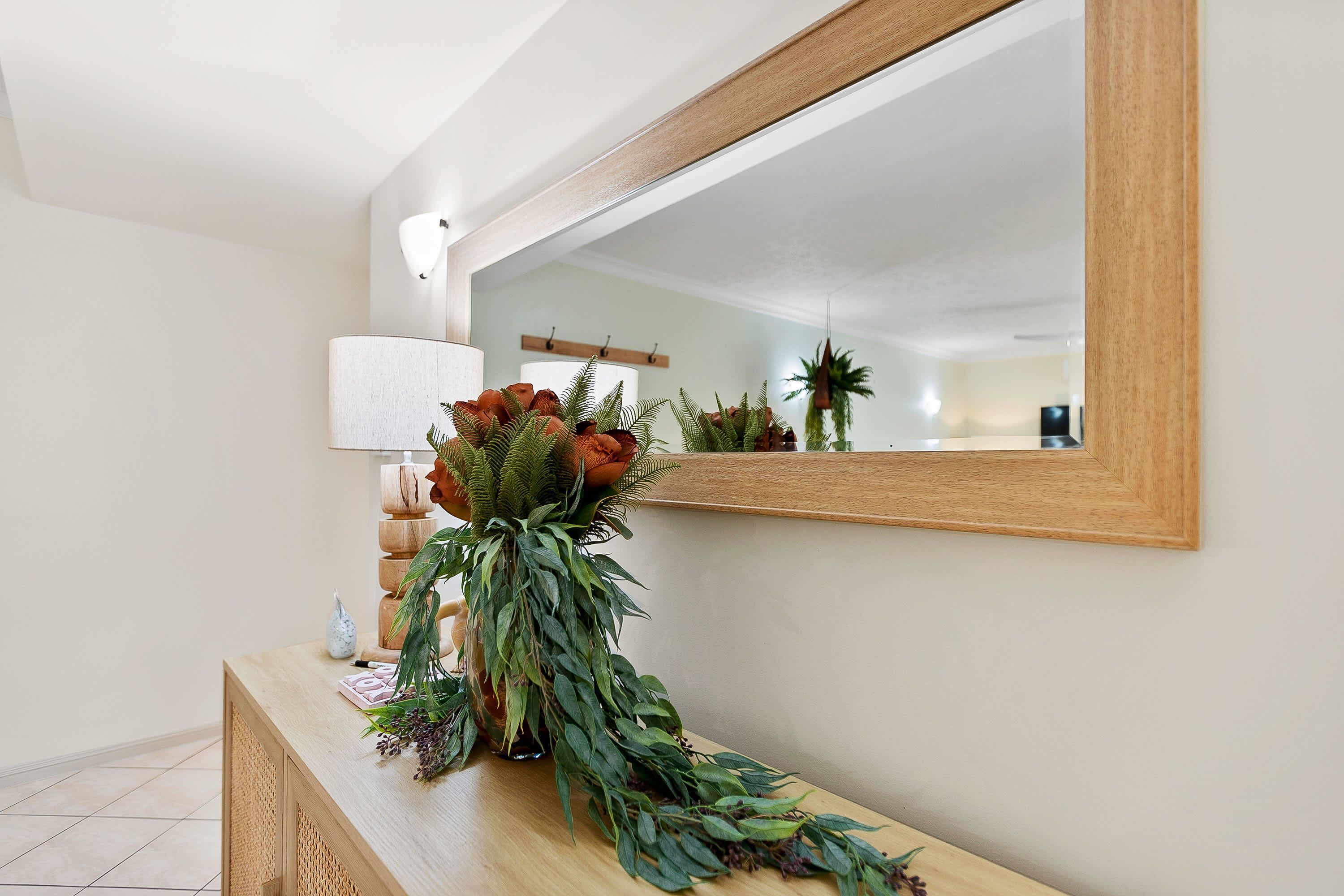 A wooden console table with a vase of green foliage and red flowers, a lamp, and a mirror on the wall.
