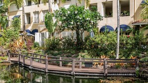 A wooden bridge over a pond with lily pads, surrounded by lush greenery and a building with balconies. - Lakeside Luxe Retreat - Pool View BBQ Spa Bath (Cairns)