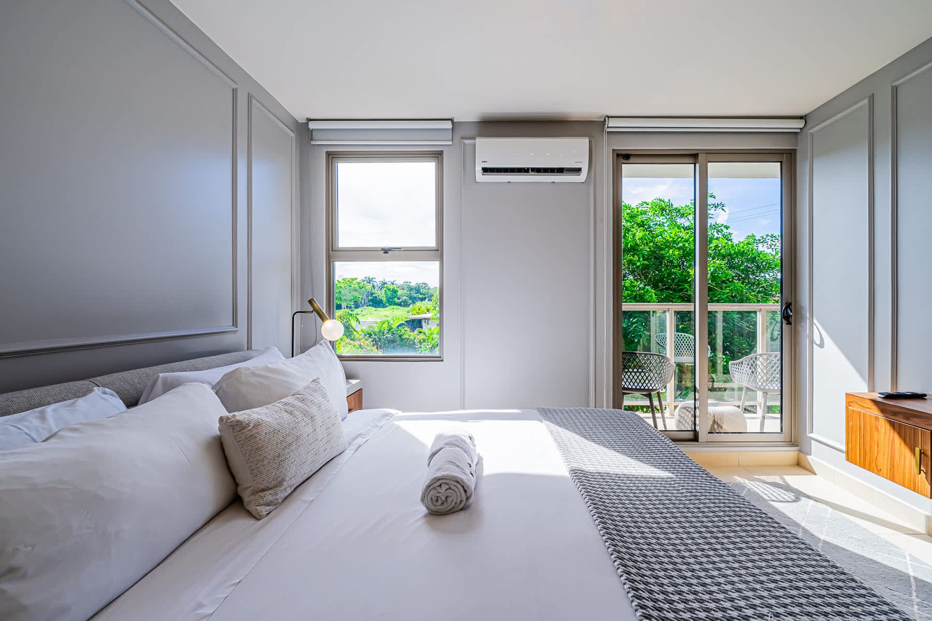 A modern bedroom with a large bed, a nightstand, an air conditioning unit, and a balcony with outdoor furniture.