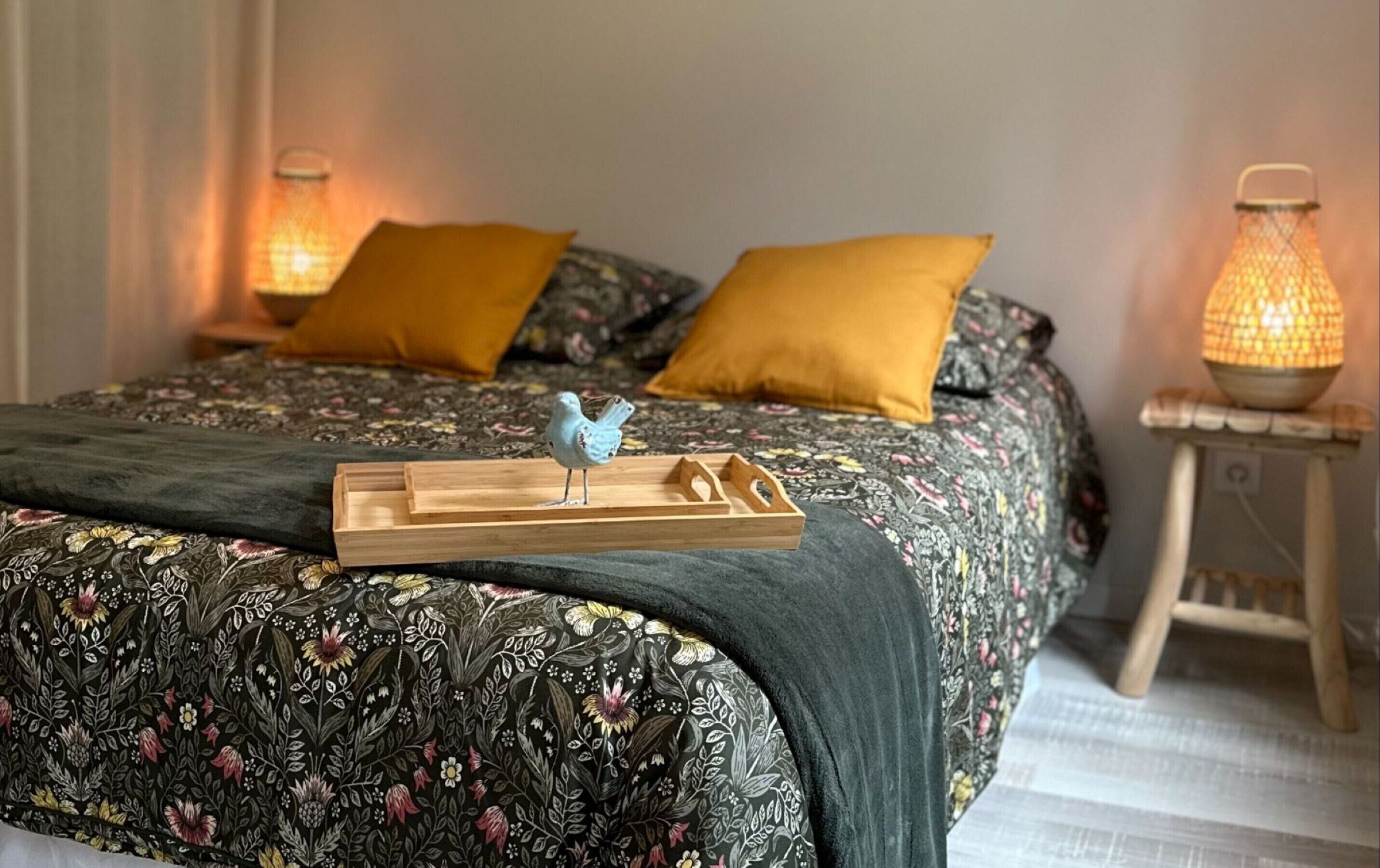 A neatly made bed with floral bedding, two pillows, a tray with a bird figurine, and two wall-mounted lamps.