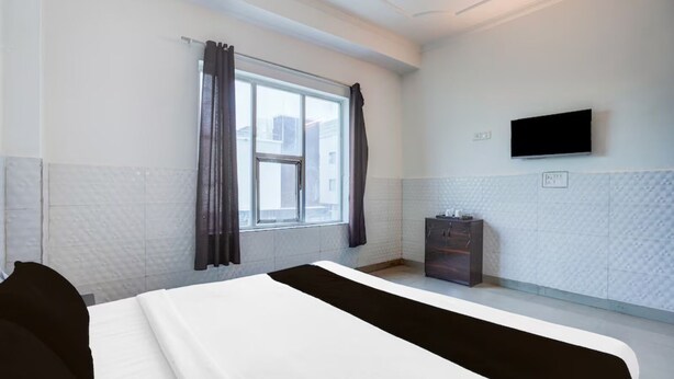 A hotel room with a bed, a window with curtains, a TV mounted on the wall, and a small cabinet. - Hotel R K Palace Agra (Agra)
