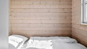 Une pièce avec un lit adossé à un mur en bois, une fenêtre sur la droite et une porte blanche sur la gauche.