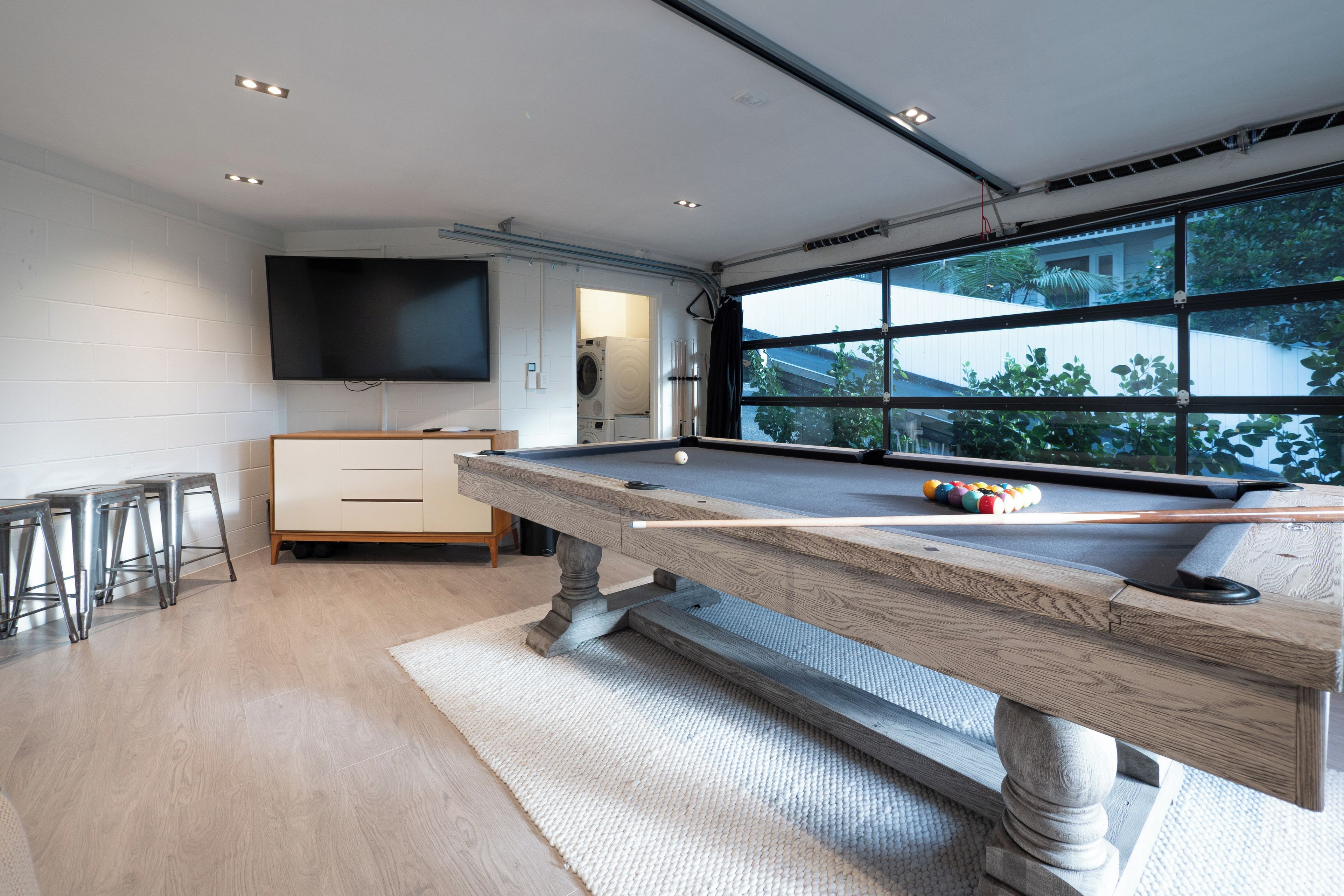 A modern living room with a pool table, a flat-screen TV, and a large window with a view of greenery.