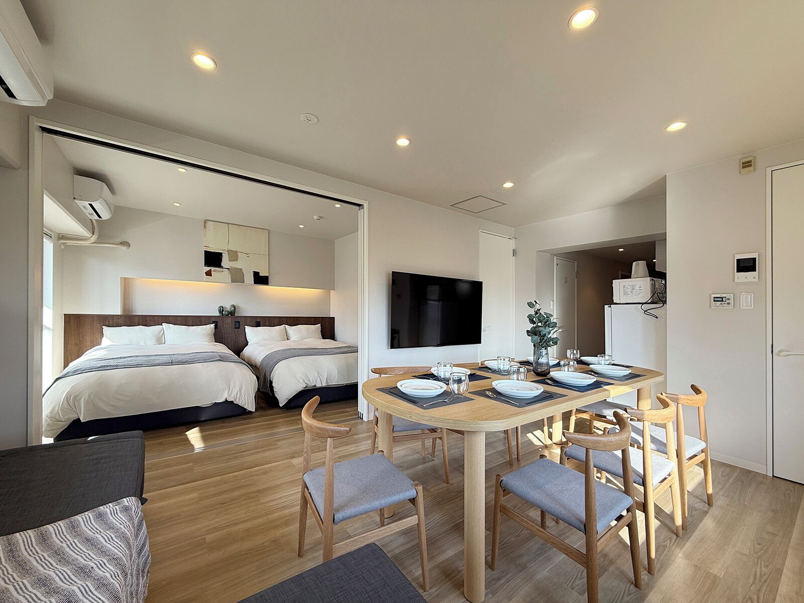 A modern hotel room with two beds, a dining area with a wooden table and chairs, and a TV mounted on the wall.