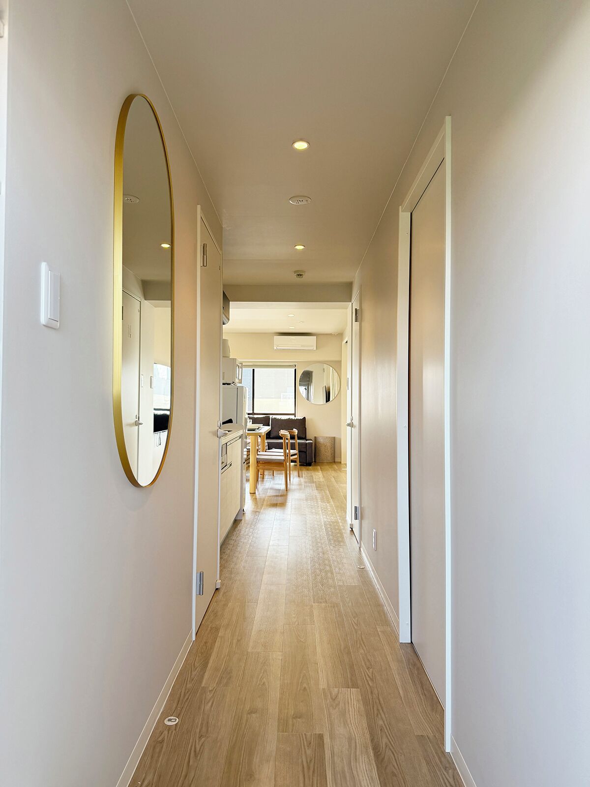 A hallway with wooden flooring, a round mirror on the left, and a kitchen area in the background.