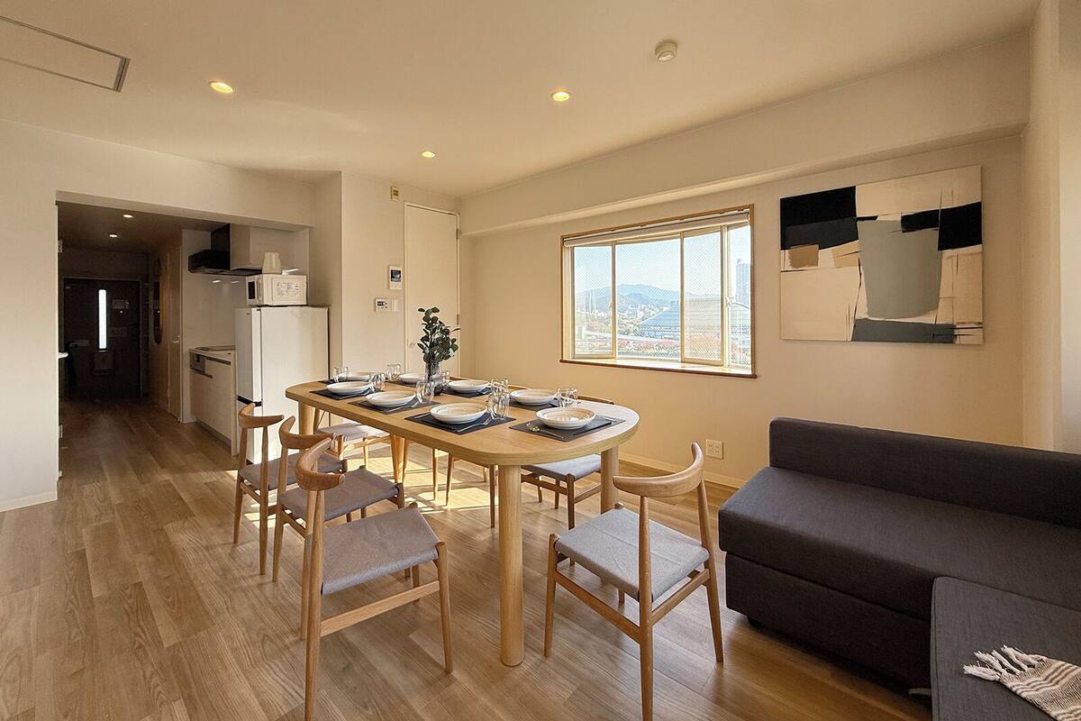 A modern dining area with a wooden table set for four, surrounded by chairs. A large window provides a view of the cityscape.