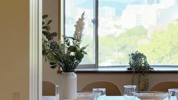 Ein Esstisch ist gedeckt mit Tellern, Gläsern und einer Vase mit Blumen, vor einem Fenster mit Blick auf die Stadt.