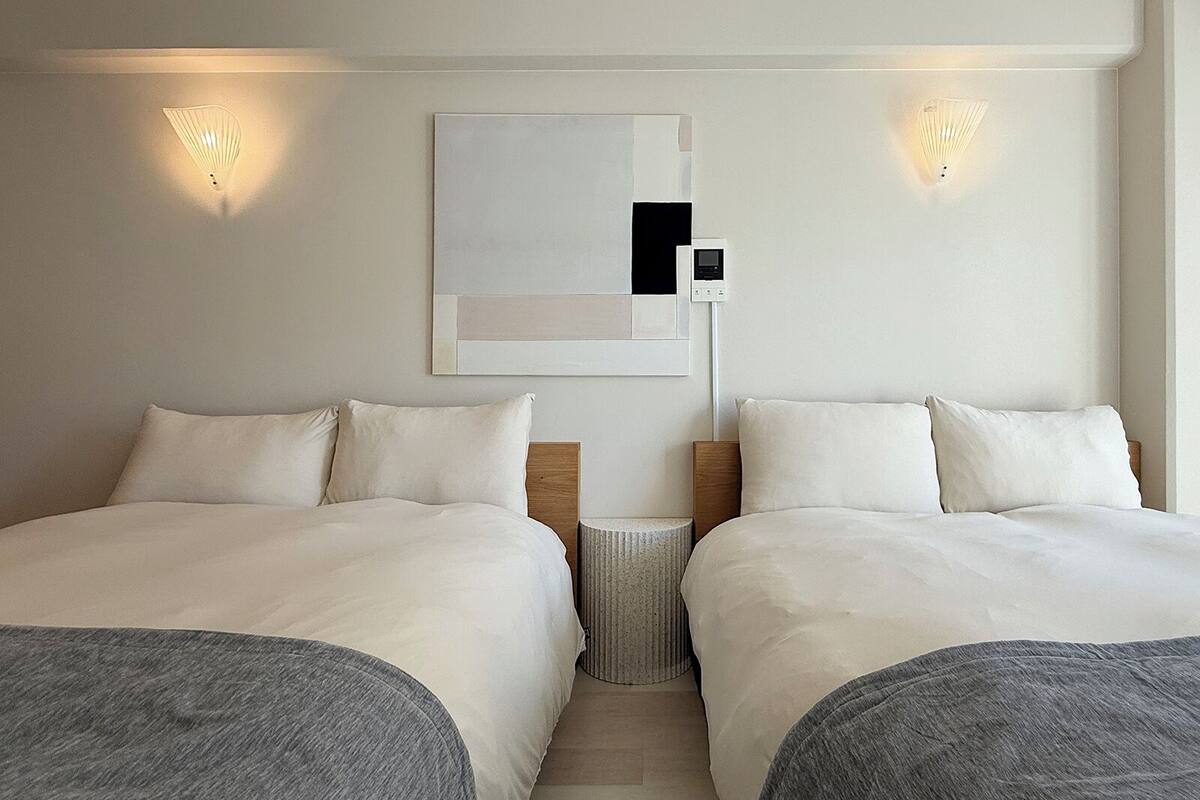 A hotel room with two beds, a small bedside table, a wall-mounted light, and a thermostat.