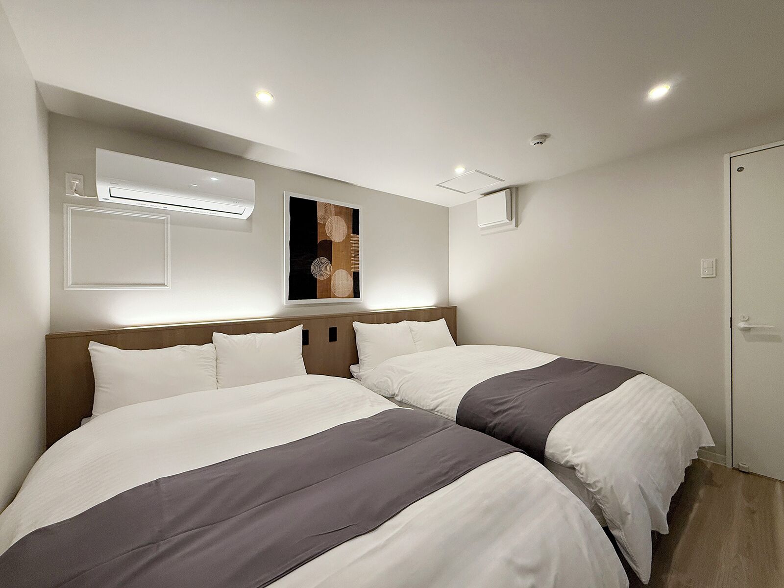A hotel room with two beds, a wooden headboard, a wall-mounted air conditioner, and a small window.
