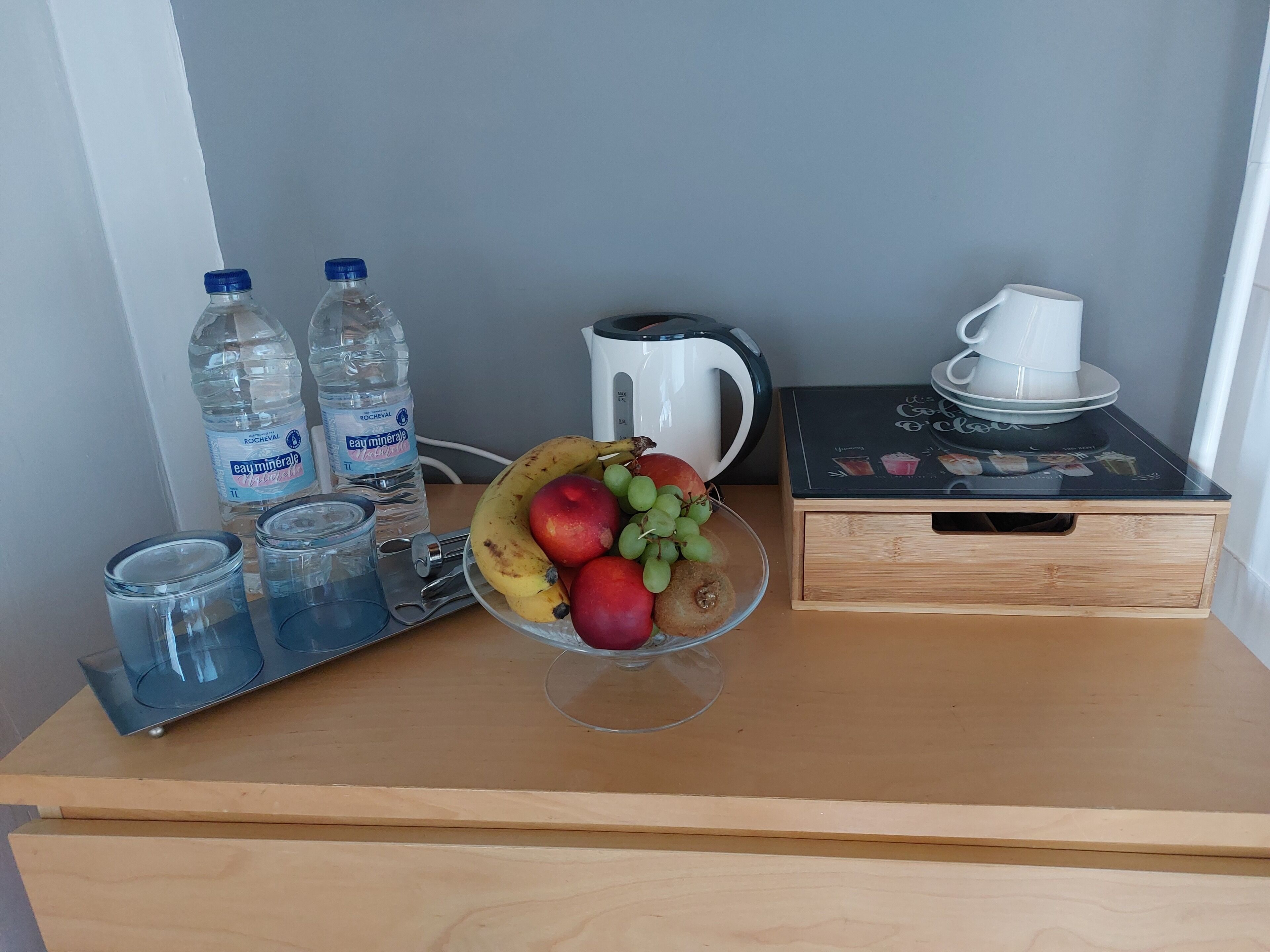 Une table en bois sur laquelle se trouvent un saladier en verre rempli de fruits, deux bouteilles d’eau, une bouilloire, une machine à café et un plateau avec des verres.