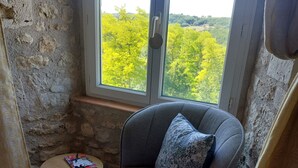 A room with a stone wall, a window with a view of green trees, a grey armchair with a patterned pillow, and a small round table with a magazine. - Charming renovated farmhouse with large pool (Lacour)