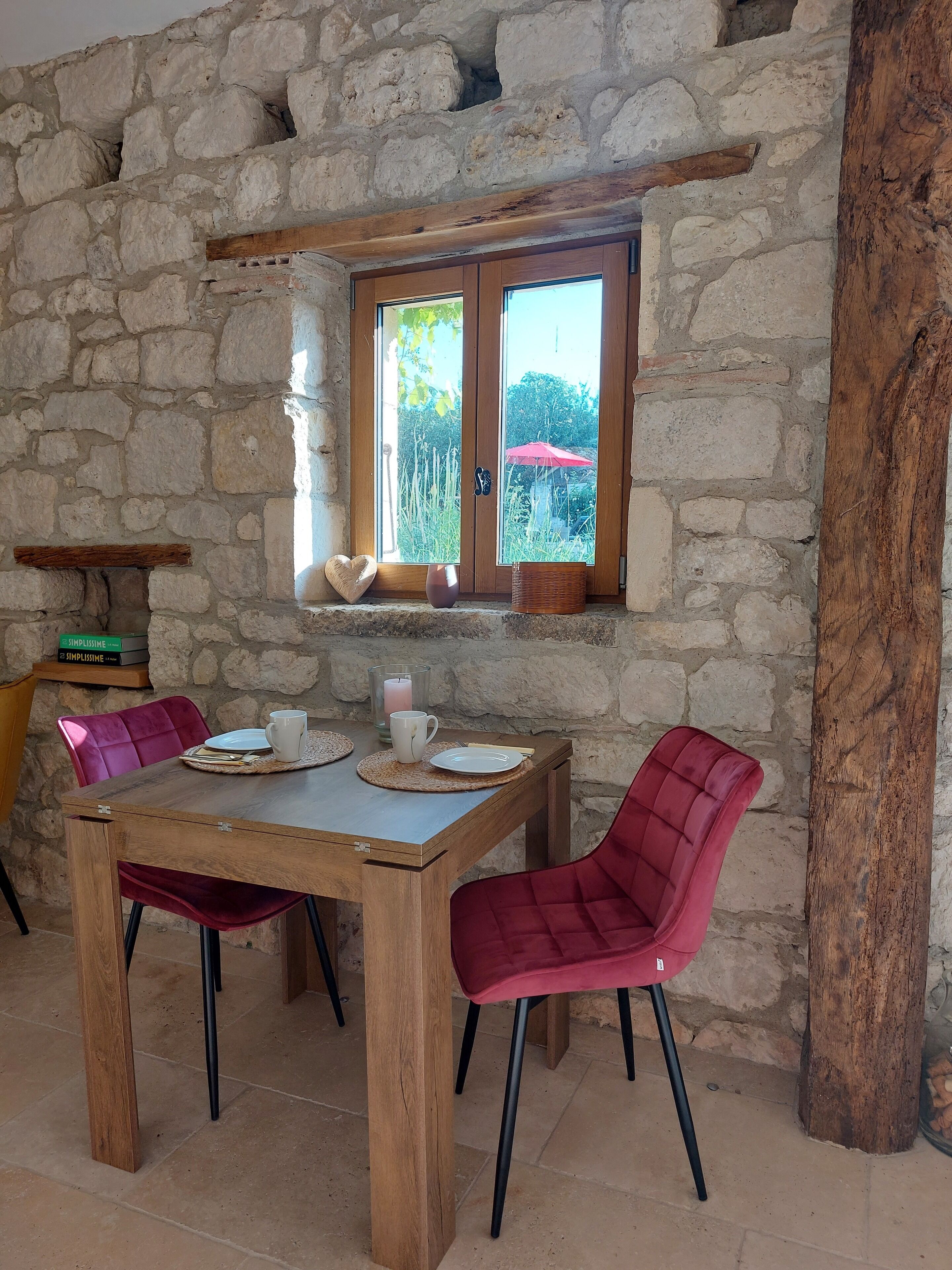 A rustic stone wall interior with a wooden table set for two, two red upholstered chairs, and a window with a view of greenery outside.
