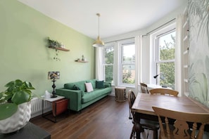 Una habitación con un sofá verde, una mesa de comedor con sillas, una lámpara, un radiador y una ventana con cortinas.