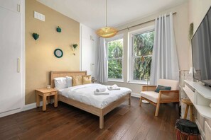 Un dormitorio con cama, mesitas de noche, una silla, televisor y ventanas grandes con cortinas.