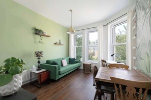 Una habitación con un sofá verde, una mesa de comedor con sillas, una lámpara, un radiador y una ventana con cortinas.