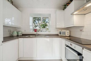 A modern kitchen with white cabinets, a built-in oven, a microwave, and a window with a view of trees. - Stylish & Elegant 1 bed flat - Watford (Hertfordshire)