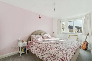 A bedroom with a bed, a nightstand, a guitar, and a window with a view of houses. - Stylish & Elegant 1 bed flat - Watford (Hertfordshire)