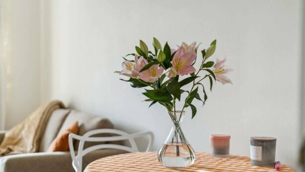 A table with a checkered tablecloth, a vase with pink flowers, and two candles on the table.
