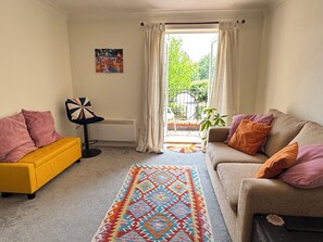 A living room with a yellow sofa, a beige sofa, and a colorful rug. There is a black chair with a geometric pattern, a radiator, and a window with curtains.