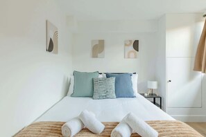 Una cama bien tendida con toallas enrolladas, una mesita de noche con lámpara y una puerta con cortina.