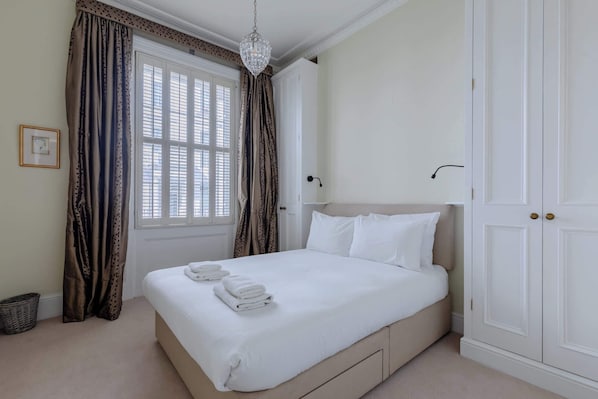 A neatly made bed with white linens, a chandelier, and a large window with curtains.