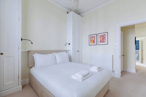 A neatly made bed with white linens and towels, a white wardrobe, and two framed artworks on the wall.