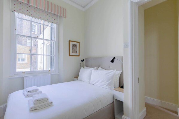 A neatly made bed with white linens and towels, a bedside table with a lamp, a window with a striped valance, and a framed picture on the wall.