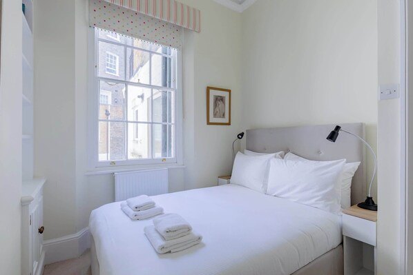 A neatly made bed with white linens and towels, a bedside table with a lamp, a window with a striped valance, and a framed picture on the wall.