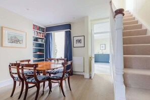 Een eethoek met een houten tafel en stoelen, een boekenkast en uitzicht op een keuken door een open deur.